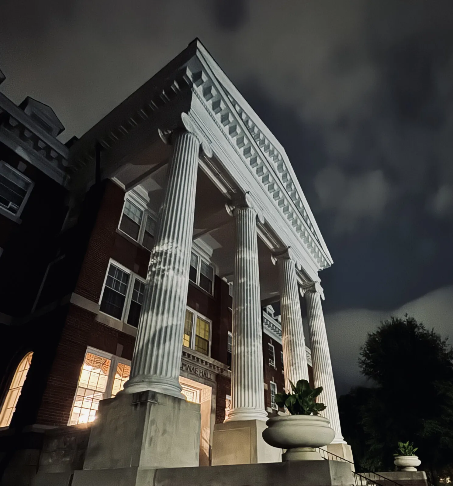 White Ionic columns of a historic brick hall glowing under floodlights against a stormy night sky