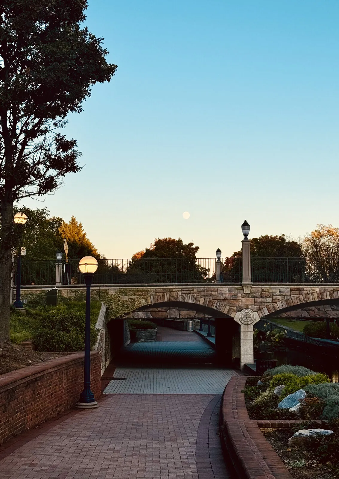 Brick walkway curving under a stone arch bridge at dusk, globe lamps lit and the moon low in a clear autumn sky