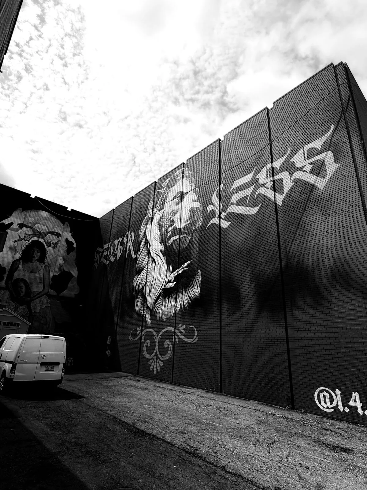 Black-and-white lion mural with 'Fear Less' lettering towering over a downtown alley under a cloud-filled sky