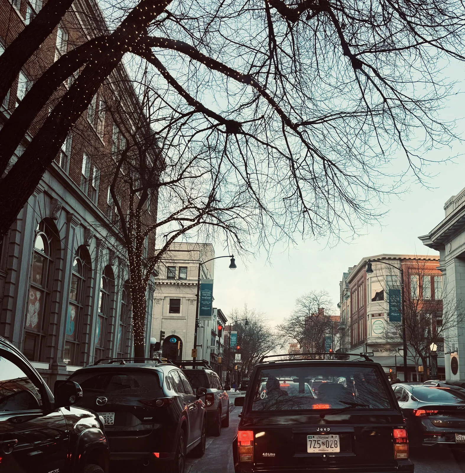 Winter dusk on a historic downtown street, bare trees wrapped in string lights above traffic waiting at the light