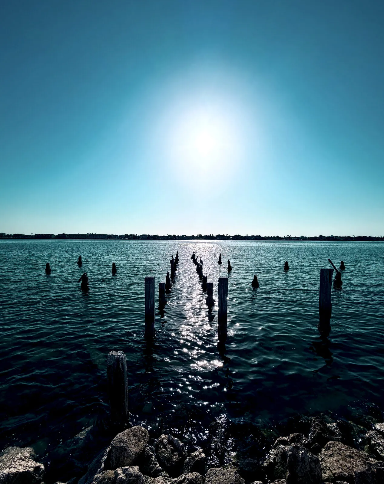 Old pilings leading toward the sun across bright coastal water