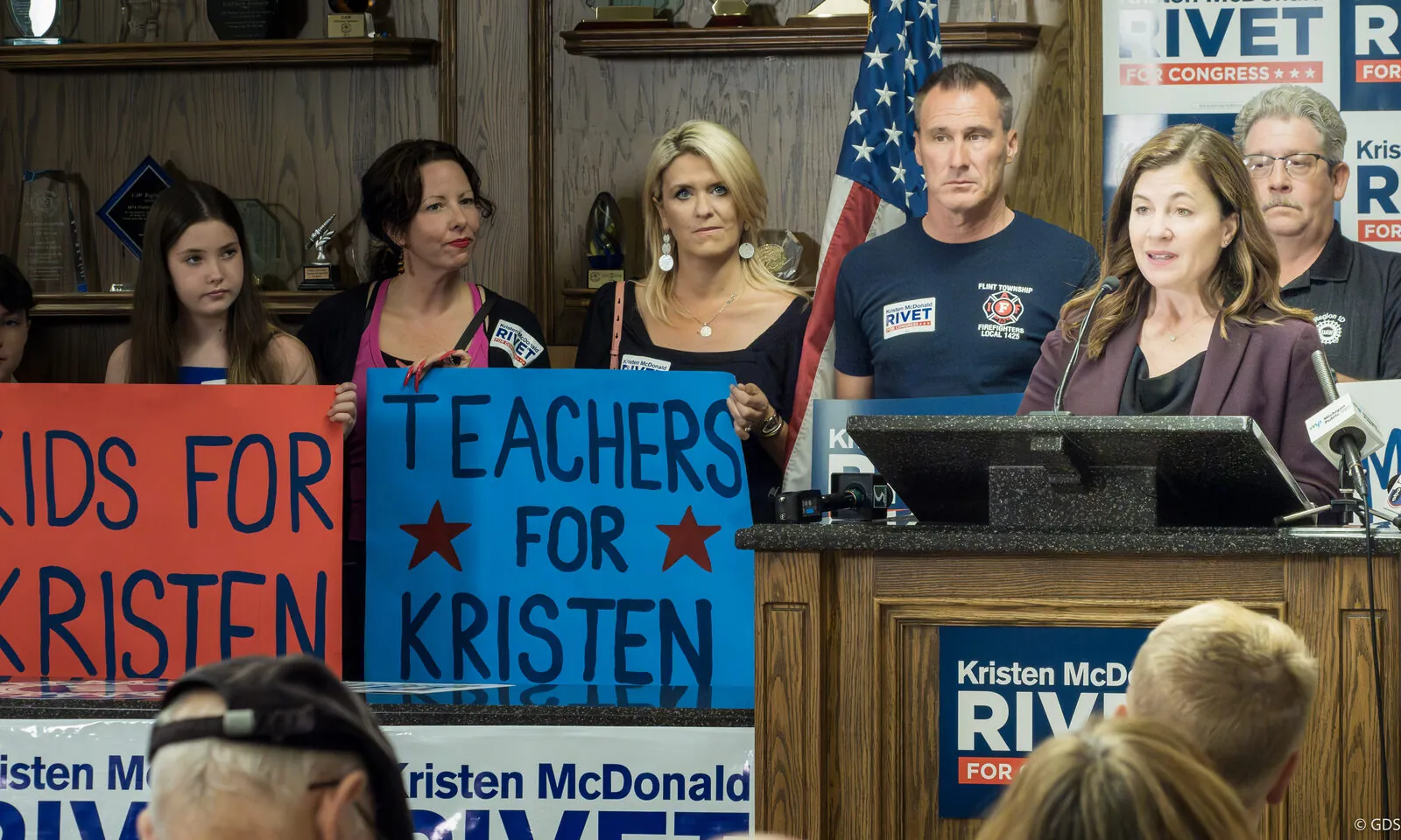 Congressional candidate Kristen McDonald Rivet speaking at a campaign event as supporters hold 'Teachers for Kristen' signs