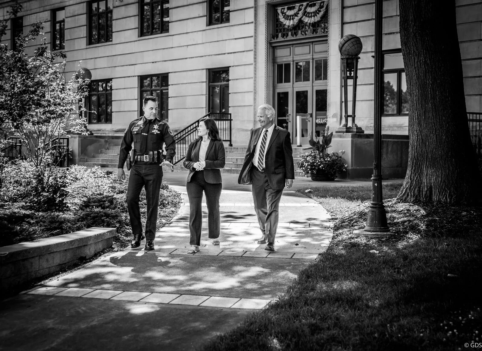 Black-and-white campaign ad shoot outside the county courthouse — a sheriff's deputy and two officials walking together