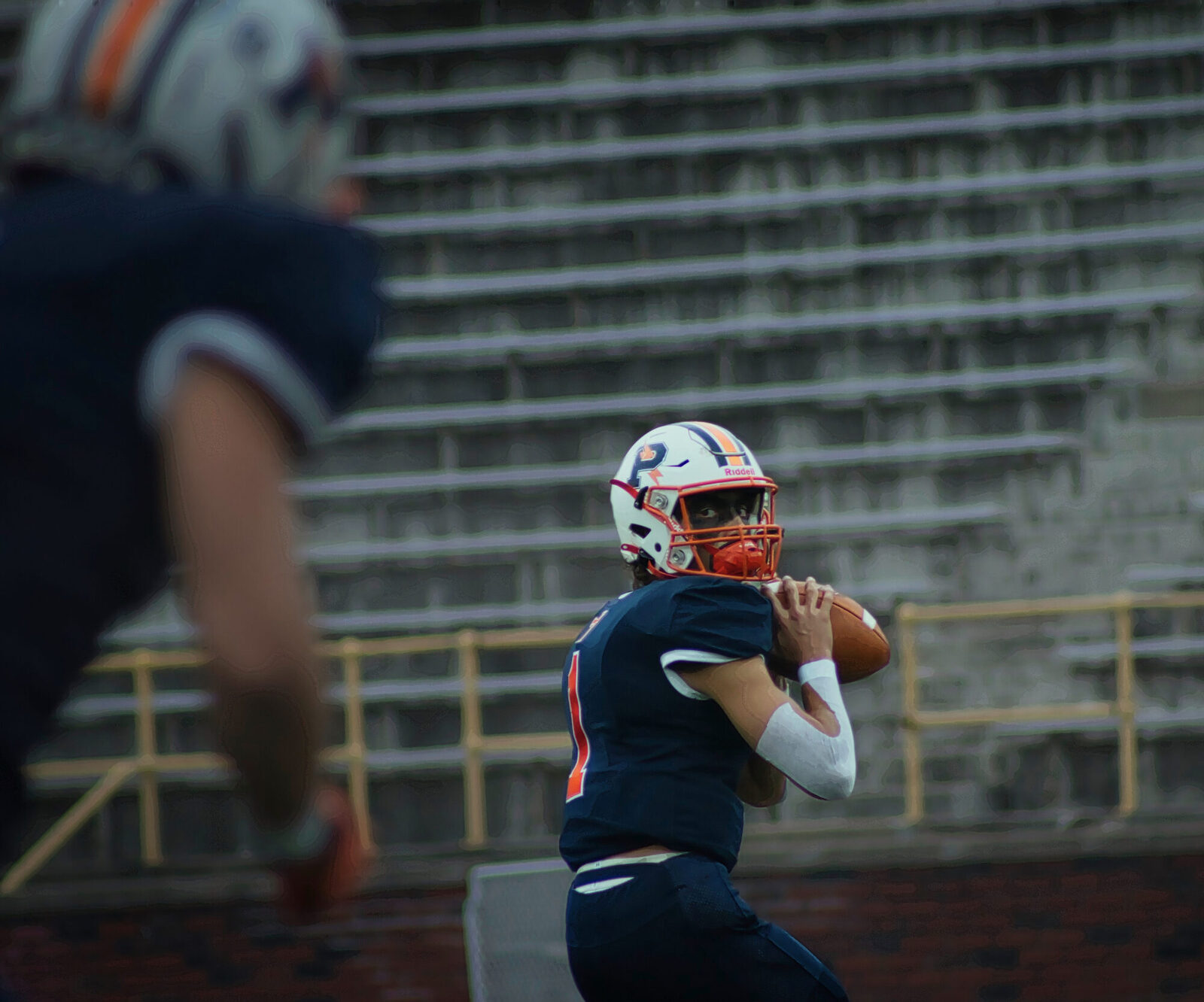 A quarterback in orange and navy sets to throw as a receiver flashes through the foreground