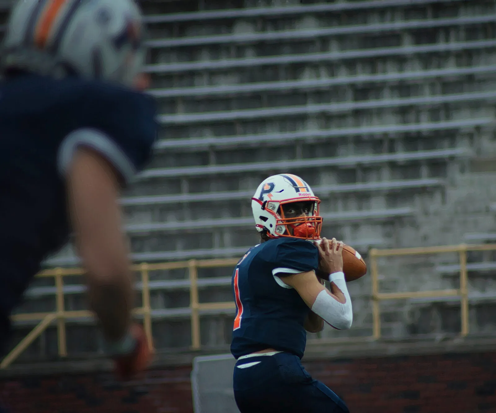 A quarterback in orange and navy sets to throw as a receiver flashes through the foreground