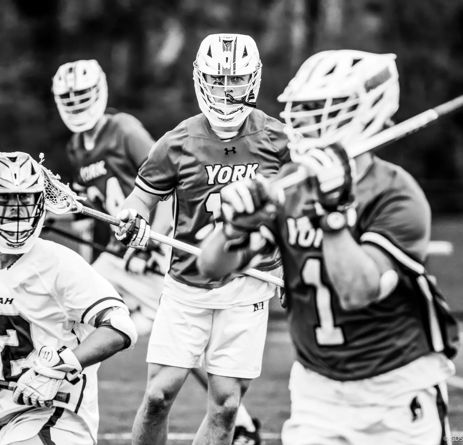 Black-and-white lacrosse — a York player stares through his facemask into the lens, teammates flanking him against Messiah