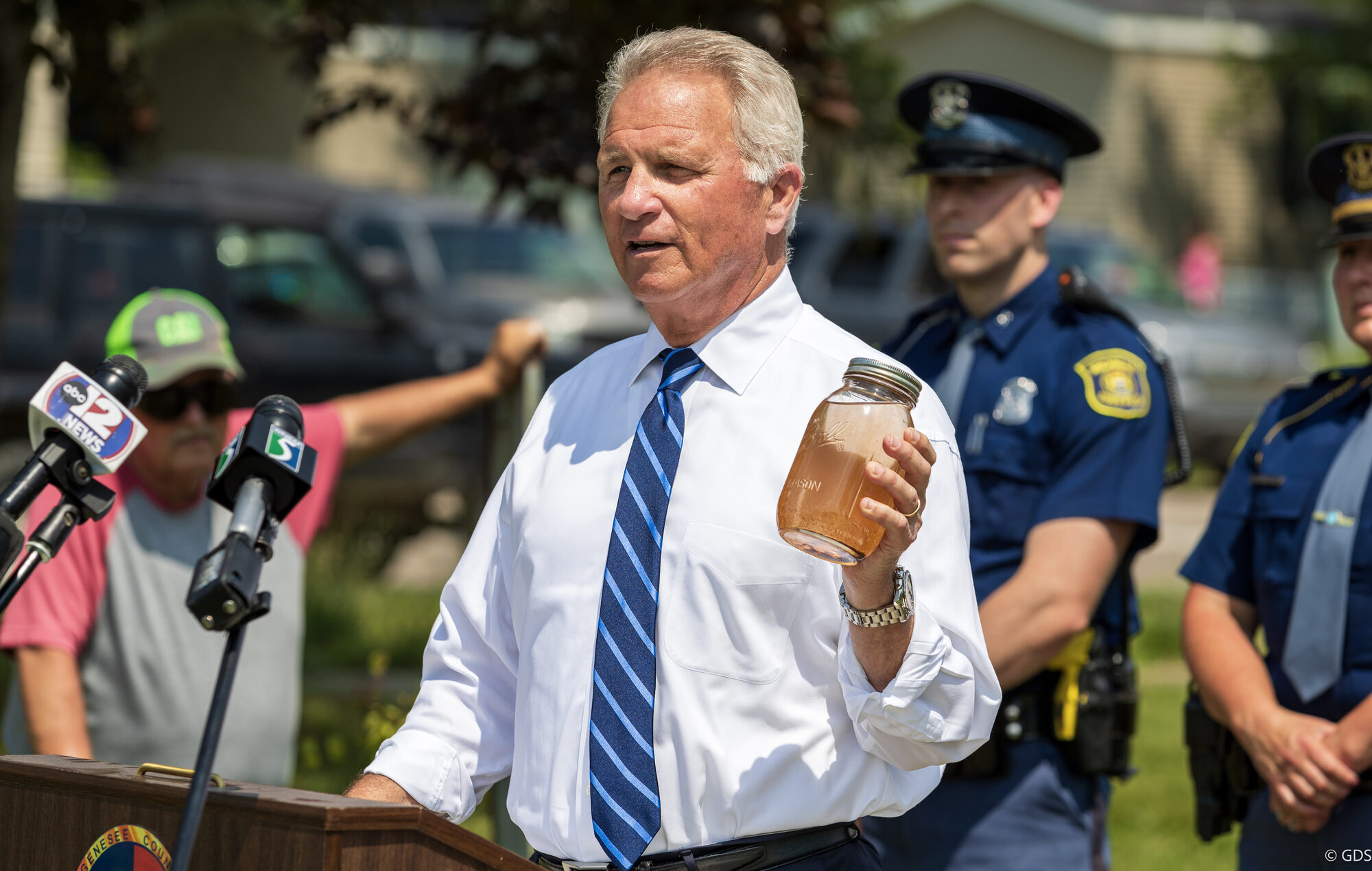 Elected official at a podium holding up a jar of discolored water at a Genesee County press conference, Michigan State Police and news microphones alongside