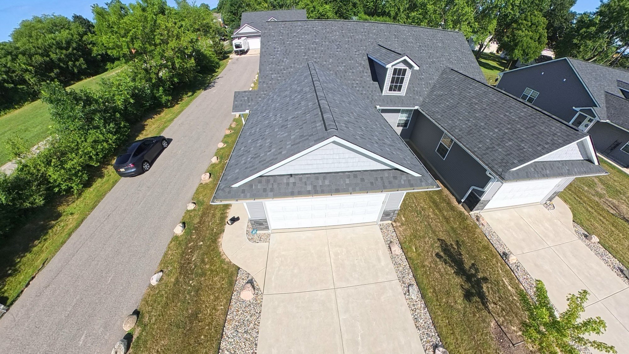 Drone view over a newly built home in Davison, gray roofline and fresh driveway leading down a quiet tree-lined street